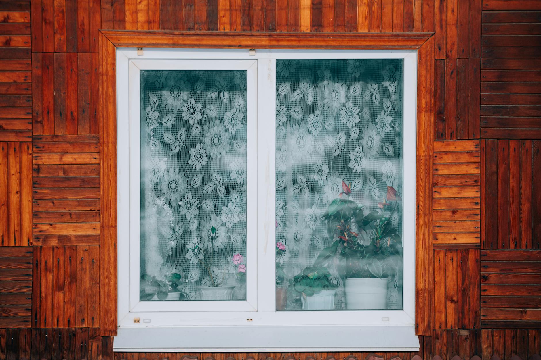 Drewniana elewacja domu z oknem i doniczkami za koronkową zasłoną